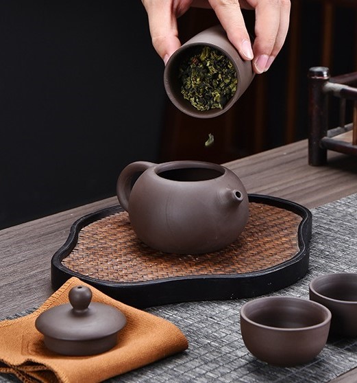 Purple clay teapot being filled with loose leaf tea on a bamboo tray during ceremony